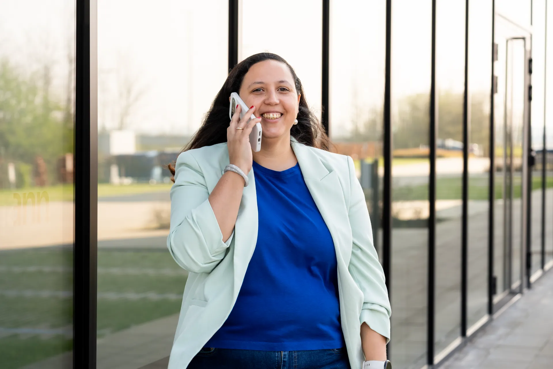 Lachende financieel adviseur Samra Daoud belt met smartphone voor modern glazen kantoorgebouw.