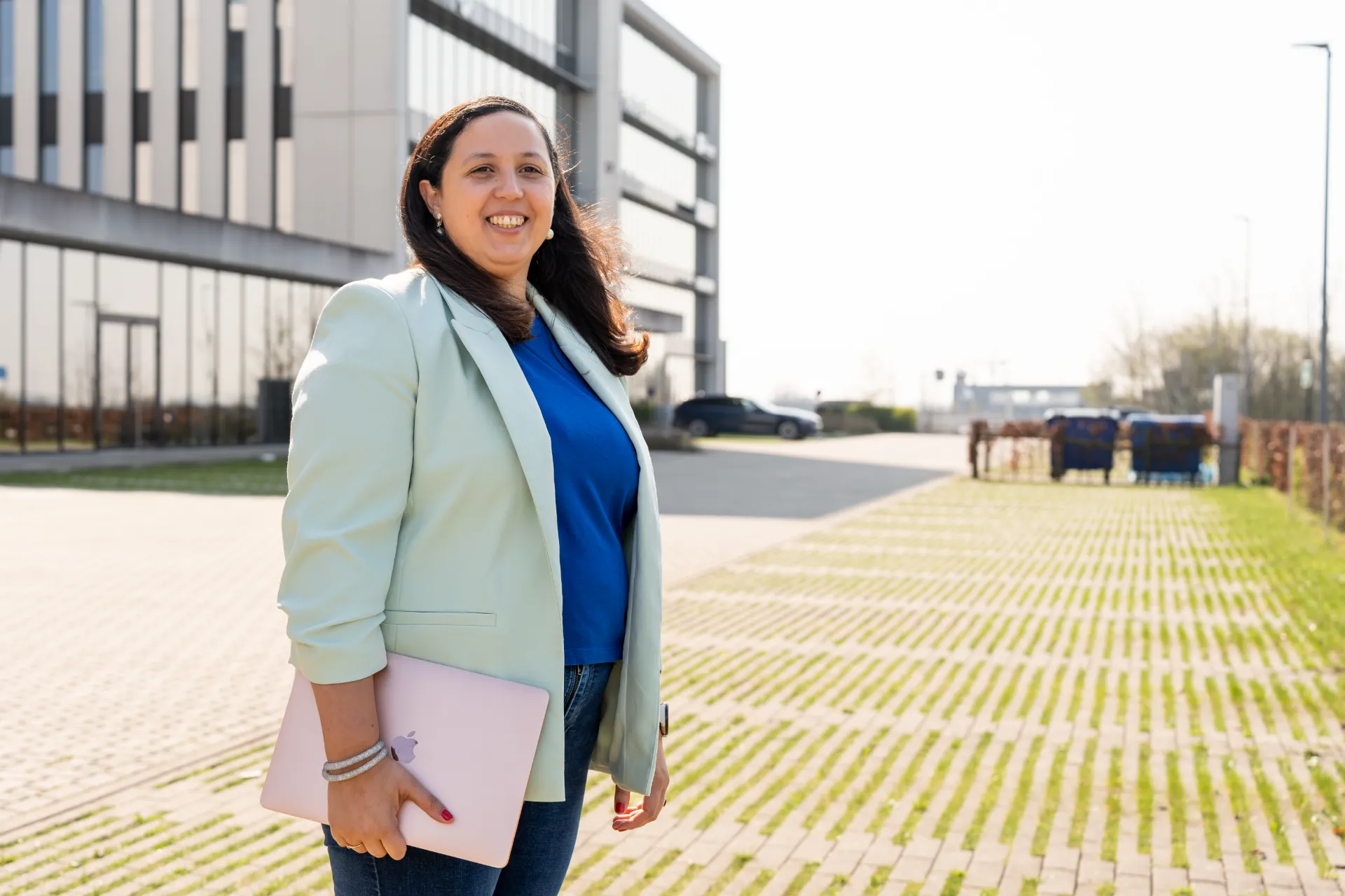 Lachende financieel adviseur Samra Daoud met roze laptop staat buiten voor een modern kantoorgebouw.