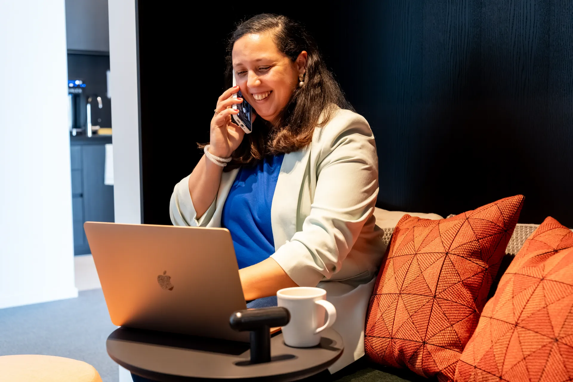 Lachende financieel adviseur Samra Daoud belt en werkt op laptop in een moderne kantooromgeving.
